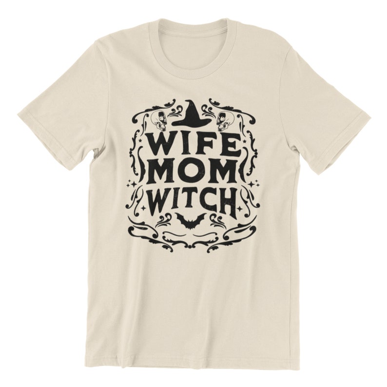 Pu&ograve; includere: Una maglietta beige con un design grafico nero che dice "Wife Mom Witch" con un cappello da strega e un pipistrello.