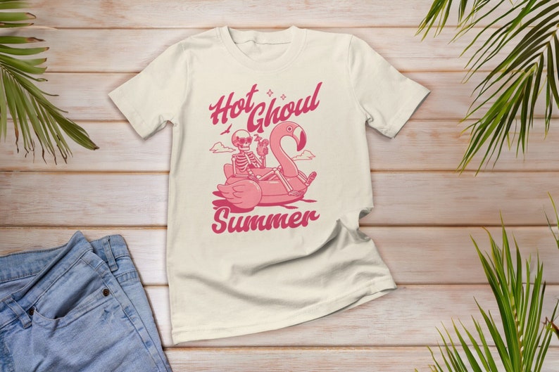 Może przedstawiać: Biały t-shirt z r&oacute;żowym nadrukiem przedstawiającym szkielet jadącego na dmuchanym flamingu z napisem "Hot Ghoul Summer".