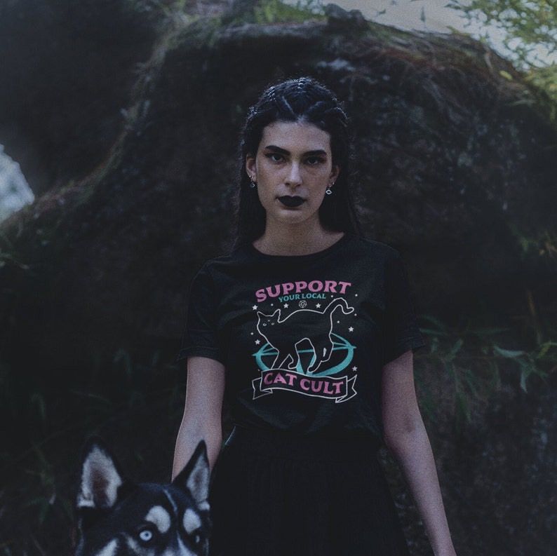 May include: A person wearing a black t-shirt with a pink and blue graphic of a black cat with the text "Support Your Local Cat Cult".