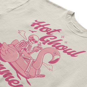 Może przedstawiać: Jasnobeżowa bluza z r&oacute;żowym nadrukiem przedstawiającym szkielet jadącego na r&oacute;żowym dmuchanym flamingu. Tekst "Hot Ghoul Summer" jest napisany r&oacute;żowym kursywą.