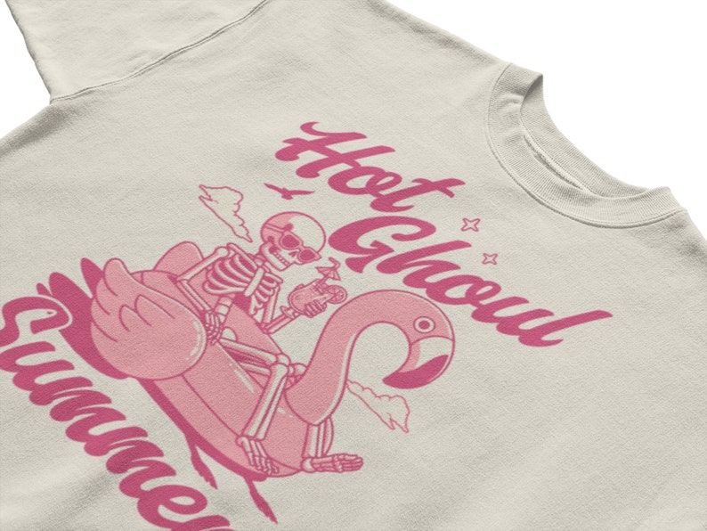Może przedstawiać: Jasnobeżowa bluza z r&oacute;żowym nadrukiem przedstawiającym szkielet jadącego na r&oacute;żowym dmuchanym flamingu. Tekst "Hot Ghoul Summer" jest napisany r&oacute;żowym kursywą.