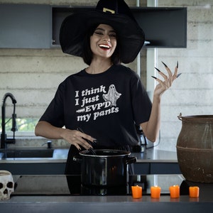 May include: A woman wearing a black witch hat and a black t-shirt with the text "I think I just EVPD'd my pants" and a ghost graphic. She is standing in a kitchen with a cauldron and candles.