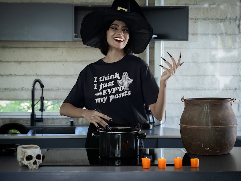May include: A woman wearing a black witch hat and a black t-shirt with the text "I think I just EVPD'd my pants" and a ghost graphic. She is standing in a kitchen with a cauldron and candles.
