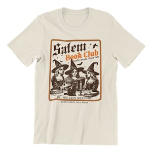 May include: A beige t-shirt with a vintage-style illustration of three witches reading books in a circle. The text reads "Salem Book Club Spells-Books-Black Cats All Witches Welcome Meets Every Full Moon".