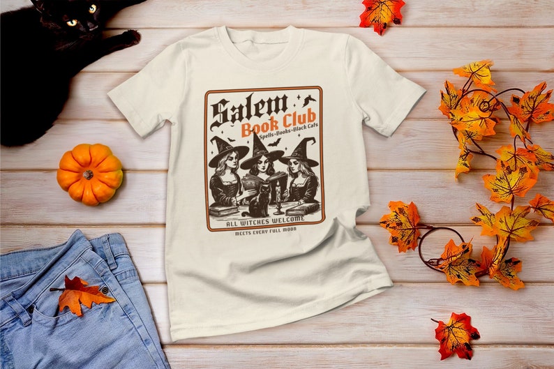 May include: A white t-shirt with a vintage-style graphic of three witches reading books in a book club. The graphic says "Salem's Book Club - Spells, Books, Black Cats" and "All Witches Welcome. Meets Every Full Moon."