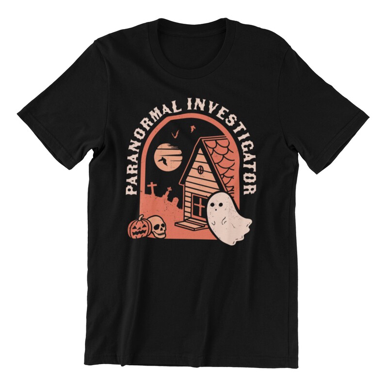 以下が含まれることがあります： 幽霊屋敷、幽霊、カボチャのグラフィックが描かれた黒いTシャツ。家の上に「Paranormal Investigator」という文字がアーチ状に書かれている。