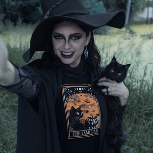 Pu&ograve; includere: Maglietta nera con grafica arancione e nera raffigurante un gatto nero, una zucca e la scritta "THE FAMILIAR". La persona indossa un cappello e un mantello da strega neri, tenendo in braccio un gatto nero.