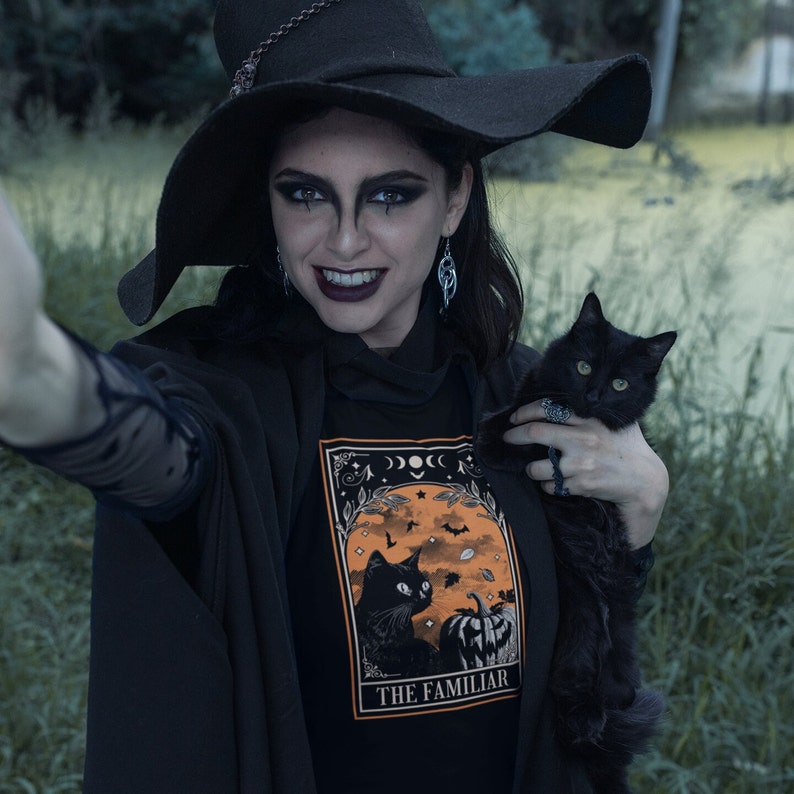 Pu&ograve; includere: Maglietta nera con grafica arancione e nera raffigurante un gatto nero, una zucca e la scritta "THE FAMILIAR". La persona indossa un cappello e un mantello da strega neri, tenendo in braccio un gatto nero.