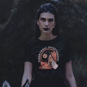 以下が含まれることがあります： 幽霊のいる家と幽霊のグラフィックが描かれた黒いTシャツ。グラフィックのテキストは「Paranormal Investigator」です。