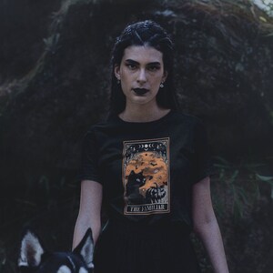 Pu&ograve; includere: T-shirt nera con grafica incorniciata raffigurante un gatto nero, zucca e fasi lunari. Il testo "THE FAMILIAR" &egrave; in basso. Lo sfondo &egrave; scuro e naturale.