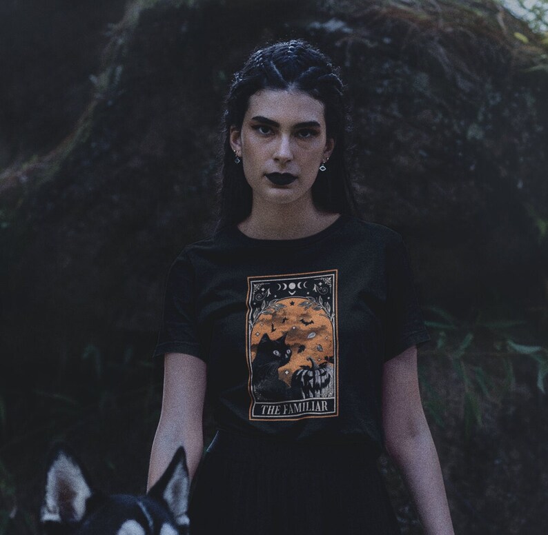 Pu&ograve; includere: T-shirt nera con grafica incorniciata raffigurante un gatto nero, zucca e fasi lunari. Il testo "THE FAMILIAR" &egrave; in basso. Lo sfondo &egrave; scuro e naturale.