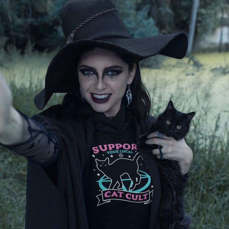 May include: A woman wearing a black witch hat and a black shirt with the text "Support Your Local Cat Cult" and a black cat in her arms.