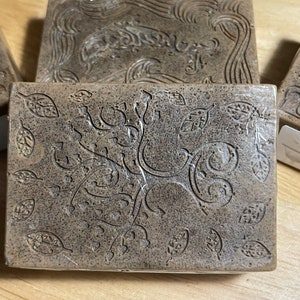 May include: Rectangular brown soap bars with intricate leaf and vine designs. The soap bars have a textured surface and are arranged on a light-colored surface. The soap bars are individually wrapped in clear plastic.