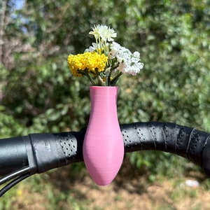 May include: A pink vase attached to a bicycle handlebar with a bouquet of yellow and white flowers.