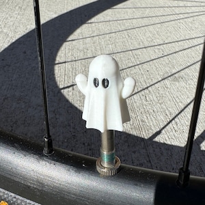 Pode incluir: Uma tampa de válvula de pneu de bicicleta em forma de fantasma branco com olhos pretos e braços estendidos. A tampa está montada num pneu de bicicleta preto com uma haste de válvula prateada. A imagem é tirada ao ar livre com luz natural.