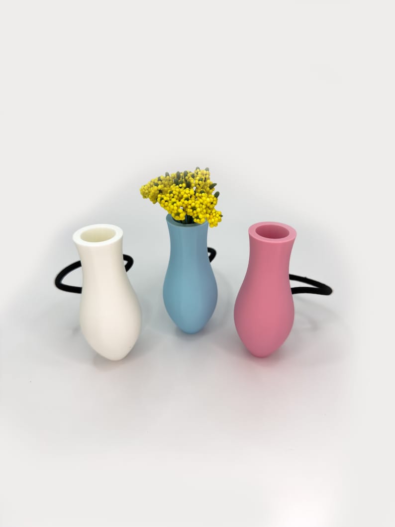 May include: Three small ceramic vases in white, light blue, and pink. The light blue vase has a small bouquet of yellow flowers.