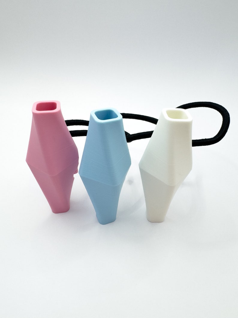 May include: Three hair ties with geometric shaped silicone ends. The hair ties are black and the silicone ends are pink, light blue, and white.