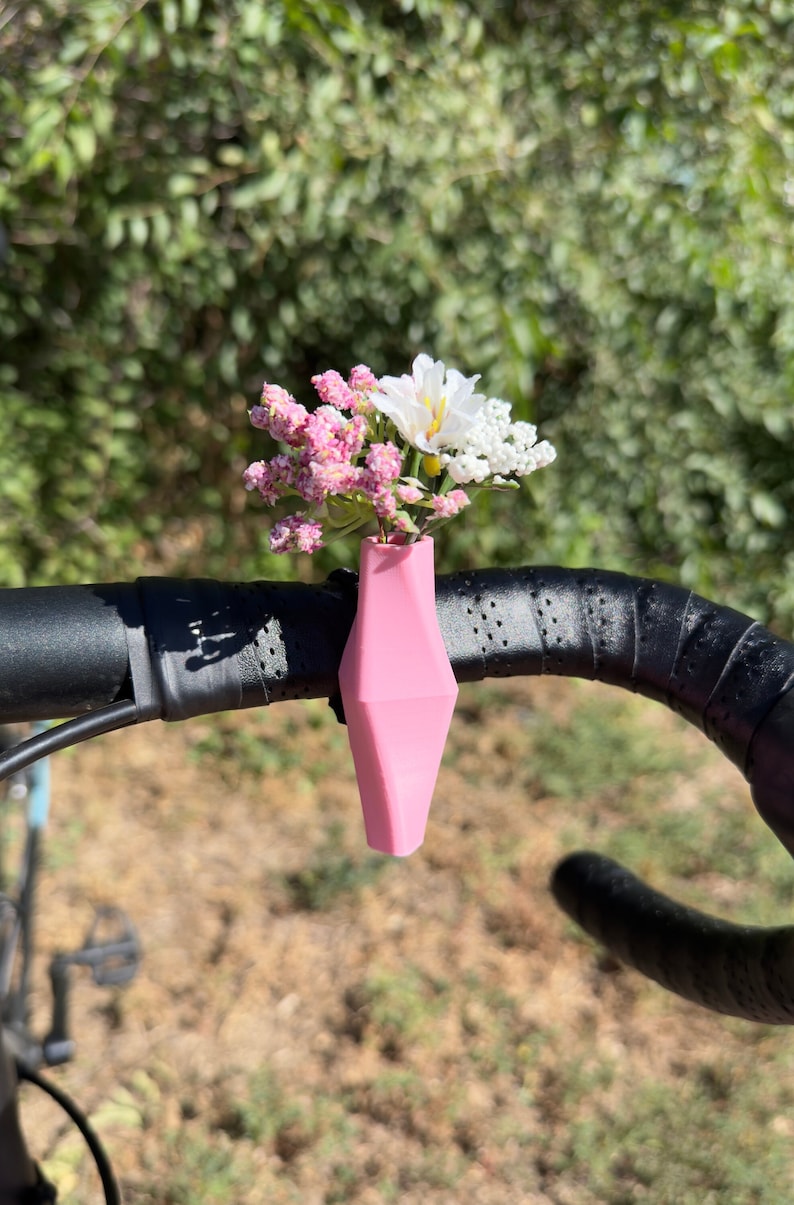 May include: A pink plastic vase attached to a bicycle handlebar with a bouquet of pink and white flowers.