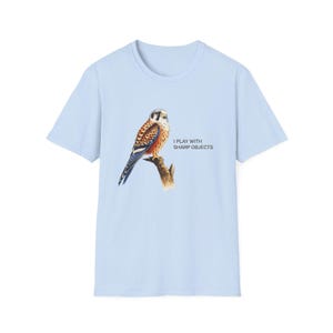 May include: A light blue t-shirt featuring a graphic illustration of a kestrel perched on a branch. To the right of the bird, the text "I PLAY WITH SHARP OBJECTS" is printed in black capital letters. The shirt has short sleeves and a crew neckline.