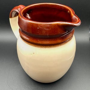 May include: A ceramic pitcher with a brown interior and rim, a tan and brown exterior, and a curved handle. The pitcher has a classic, rounded shape and a spout for pouring.