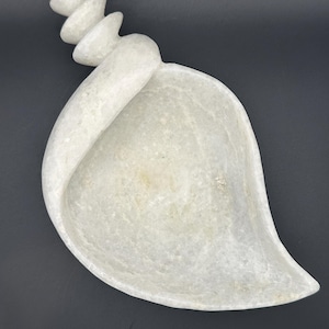 May include: A light-colored, leaf-shaped decorative dish with a curved handle and a stacked, tiered design at the top. The dish is made of a stone-like material and has a smooth, polished surface. The background is dark.