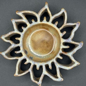 May include: A ceramic sun-shaped dish with a central bowl and petal-like edges. The dish has a brown and tan glaze with hints of blue. The design is decorative and suitable for holding small items or as a decorative piece.