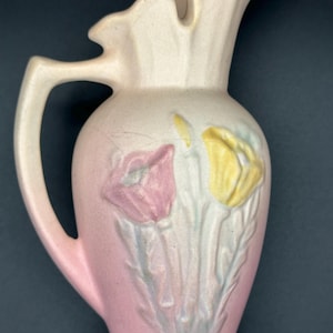 May include: A vintage ceramic pitcher in shades of pink and cream, featuring embossed floral designs. The pitcher has a ruffled top and a curved handle. The flowers are pink and yellow, with green stems. The pitcher is approximately 8 inches tall.