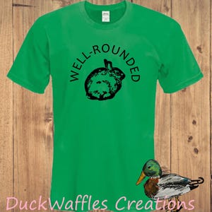 May include: A bright green short-sleeved t-shirt featuring a black graphic of a round rabbit in the center. Above the rabbit, the text WELL-ROUNDED is printed in black. In the bottom right corner, the text DuckWaffles Creations is visible.