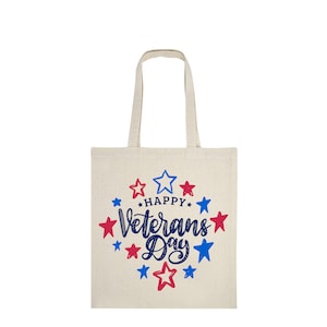 Peut inclure: Un sac fourre-tout en toile beige avec l'inscription "Happy Veterans Day" en bleu, entour&eacute;e d'&eacute;toiles rouges et bleues. Le sac poss&egrave;de deux anses et est con&ccedil;u pour transporter des objets.