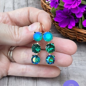Könnte beinhalten: Ein Paar goldfarbene Ohrhänger mit drei irisierenden, smaragdgrünen und türkisfarbenen Kristallsteinen. Die Ohrringe werden von einem goldfarbenen Haken gehalten. Die Ohrringe sind von Sparkled Rose Jewelry.