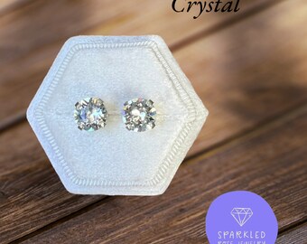 May include: A pair of sparkling crystal stud earrings displayed in a white velvet hexagonal box. The word "Crystal" is written in cursive above the earrings. The earrings are set against a wooden background. The logo for Sparkled Rose Jewelry is in the bottom right corner.