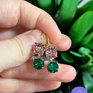 Puede incluir: Pendientes dorados con piedras preciosas rosas y verdes. Cada pendiente tiene una piedra rosa redonda sobre una piedra verde cuadrada. Los pendientes se sostienen en una mano, con un fondo borroso de plantas verdes.