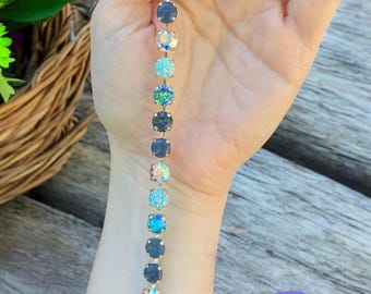 May include: A silver bracelet with a series of round, iridescent and blue-toned crystals. The bracelet is held up against a wooden surface. The bracelet is from Sparkled Rose Jewelry.