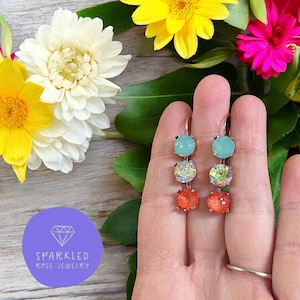 May include: A pair of dangle earrings with three colorful, round stones. The top stone is light blue, the middle is iridescent, and the bottom is orange. The earrings are held up by a hand, with flowers in the background.