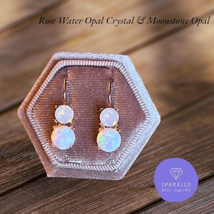 Pode incluir: Brincos em ouro rosa com duas pedras de opala brancas. Os brincos são exibidos numa caixa de joias de veludo. O texto "Rose Water Opal Crystal & Moonstone Opal" está na imagem. Da Sparkled Rose Jewelry.