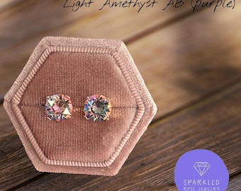 May include: Two round, faceted Light Amethyst AB purple stud earrings with an iridescent finish, displayed in a hexagonal, dusty rose velvet jewelry box. The text Light Amethyst AB (purple) is visible above the earrings.