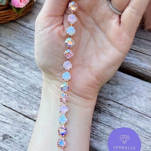 Pode incluir: Uma pulseira em ouro rosa com cristais redondos e iridescentes. Os cristais alternam entre as cores rosa, branco e opalescente. A pulseira é exibida sobre uma superfície de madeira, com uma cesta de rosas cor-de-rosa ao fundo. A pulseira está rotulada como "Sparkled Rose Jewelry."