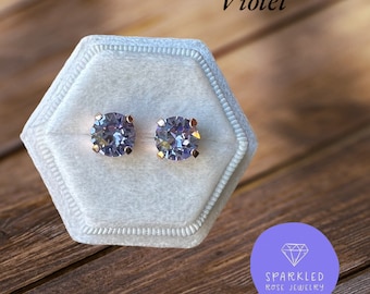 May include: A pair of violet-colored stud earrings with a round, faceted design, set in a rose gold metal. The earrings are displayed in a light gray velvet jewelry box. The word "Violet" is written in a script font.