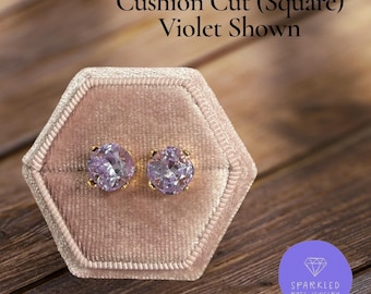 May include: A pair of violet cushion-cut stud earrings with gold settings, displayed in a pink velvet jewelry box. The text reads "Cushion Cut (Square) Violet Shown". The earrings are from Sparkled Rose Jewelry.