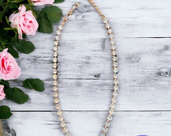Puede incluir: Un collar dorado con cristales iridiscentes, exhibido sobre una superficie de madera blanca. El collar está adornado con numerosas piedras redondas y brillantes. También se ven rosas rosas y el texto "Sparkled Rose Jewelry".