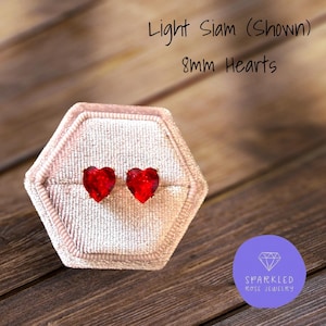 May include: Two 8mm heart-shaped red earrings presented in a pink hexagonal box. The text "Light Siam (Shown)" and "8mm Hearts" is above the earrings. The Sparkled Rose Jewelry logo is in the lower right corner.