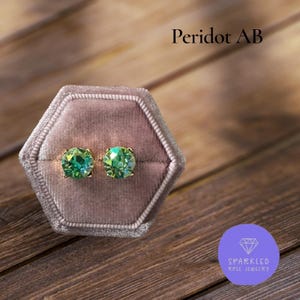 May include: Two peridot AB stud earrings in a pink velvet box. The earrings have a gold setting and feature round, faceted green gemstones. The text "Peridot AB" is above the box, and a logo for "Sparkled Rose Jewelry" is below.