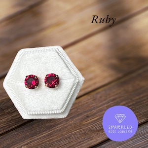 May include: A pair of ruby-colored stud earrings with gold-colored posts, displayed in a white velvet jewelry box. The word "Ruby" is written in black script above the earrings. The earrings are from Sparkled Rose Jewelry.