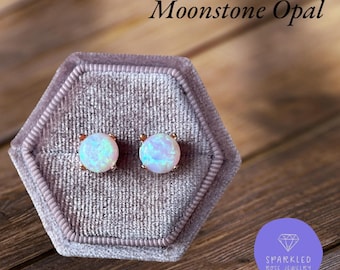 Puede incluir: Un par de pendientes de botón redondos de ópalo de piedra lunar con engaste de oro rosa, presentados en una caja de joyería de terciopelo gris. Los pendientes tienen un brillo nacarado e iridiscente. El texto "Moonstone Opal" está arriba.