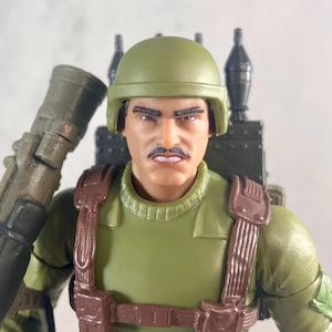 May include: A close-up of a military action figure. The figure is wearing a green uniform, helmet, and brown straps. It has a mustache and is holding a weapon. A backpack with missiles is visible.