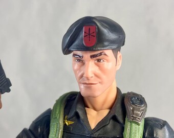 May include: A detailed action figure of a man in a black uniform and beret. The figure has a green harness with gold-colored details and a belt with gold-colored bullets. The beret features a red emblem.