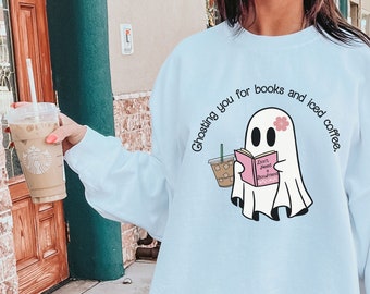 Puede incluir: Sudadera blanca con un diseño de fantasma sosteniendo un libro y una taza de café. El texto en la sudadera dice "Ghosting you for books and iced coffee". El fantasma tiene una flor rosa. La sudadera se usa con zapatillas blancas.