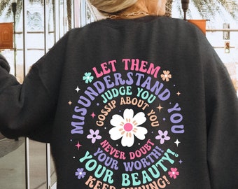 Op de afbeelding: Een donkergrijs sweatshirt met een kleurrijke grafische print die zegt "Laat ze je misverstaan, beoordelen, roddelen over je. Twijfel nooit aan je waarde, je schoonheid! Blijf stralen!"