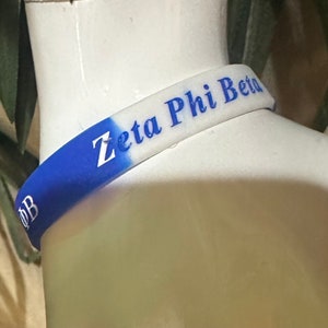 Puede incluir: Una pulsera de silicona azul y blanca con el texto "Zeta Phi Beta" en letras azules.