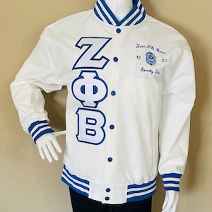 Può includere: Giacca college bianca con dettagli blu. Presenta le lettere greche ZΦB in blu. La giacca ha strisce blu sul colletto, polsini e orlo. Il testo "Zeta Phi Beta Sorority Inc." è ricamato sul petto destro.